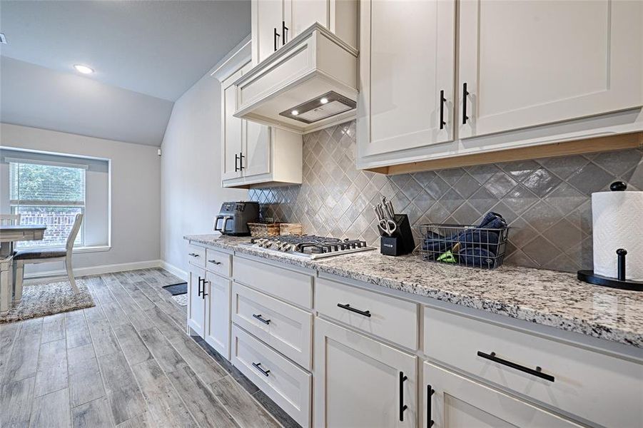 Kitchen with white cabinets, backsplash, light hardwood / wood-style floors, stainless steel gas stovetop, and custom range hood