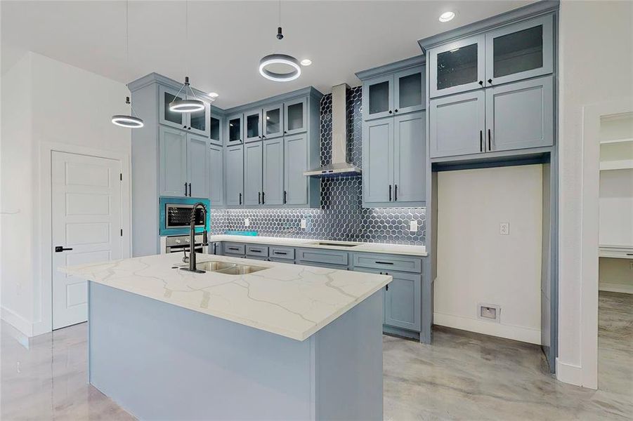 Kitchen with wall chimney range hood, a kitchen island with sink, light stone countertops, decorative backsplash, and decorative light fixtures