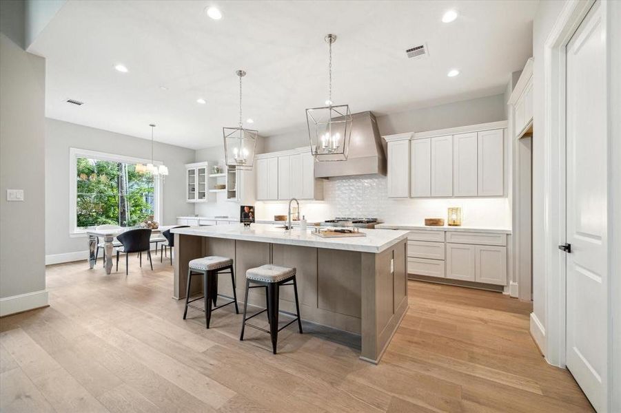 This is a spacious, gourmet kitchen with white cabinetry, stainless steel appliances, a central island with bar seating, and elegant pendant lighting. Natural light flows in through a large window overlooking greenery. The kitchen opens to a dining area, perfect for entertaining.